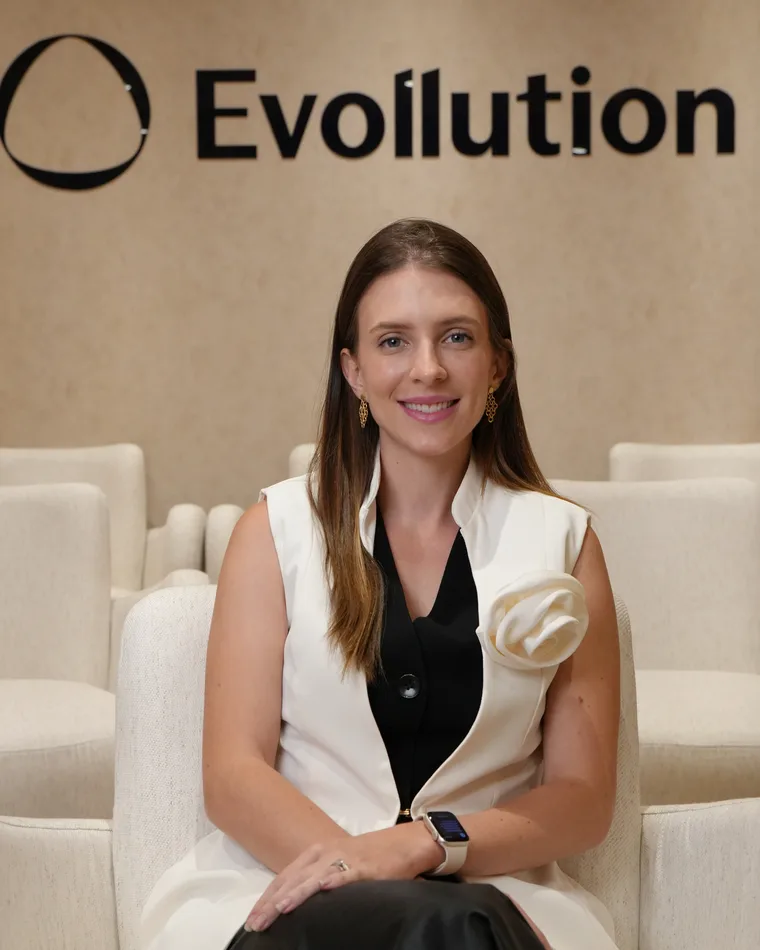 Dra. Thaís Lancaster sentada em poltrona clara, com o painel Evollution ao fundo